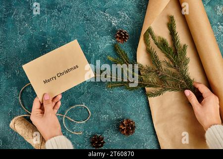 Zugeschnittene Ansicht einer Frau mit Merry Christmas Umschlag und Kiefernzweig in der Nähe von Bastelpapier und Garn, diy Stockfoto