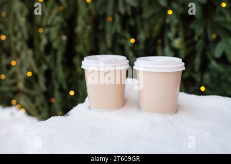 Zwei Plastikbecher stehen auf dem Schnee vor dem Hintergrund einer grünen Wand eines Weihnachtsbaums und Bokeh von einer Girlande. Kaffee zum Mitnehmen, ein Spaziergang in den fres Stockfoto