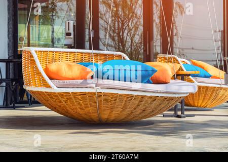 Farbenfrohe Kissen auf einem modernen Tagesbett. Braune Rattan-Liegestühle mit orangefarbenen und blauen Kissen, die mit Seilen auf der entspannenden Terrasse hängen. Sonnenliege zum Sonnenbaden und Stockfoto