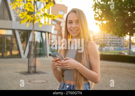 Eine fröhliche blonde Dame mit langen, lockeren Haaren lächelt in die Kamera. Die junge Frau lacht fröhlich, wenn sie Zeit im Sommer-Stadtpark verbringt Stockfoto