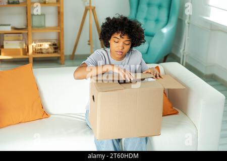Afrikanisches Mädchen, das die Lieferung auspackt und in die Schachtel schaut. Glückliche Frau, die eine Schachtel öffnet. Eine Frau bekommt ein Paket, das sich die gelieferten Waren ansieht. Zufriedene Kunden positives Feedback, schnelle Expresslieferung Stockfoto