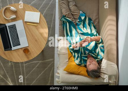 Ältere Frau, die sich auf der Couch entspannt, ihrem Freund SMS schreibt und in sozialen Medien scrollt, Blick von oben Stockfoto