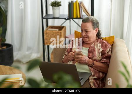 Eine ältere Frau, die auf der Couch ausruht, Tee trinkt und eine Show auf dem Laptop ansieht Stockfoto