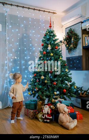 Das kleine Mädchen steht neben einem glitzernden Weihnachtsbaum und berührt die Zweige. Seitenansicht Stockfoto
