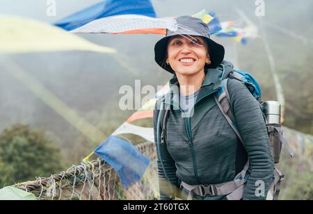 Junge lächelnde Backpackerin überquert den Canyon über die Hängebrücke mit bunten tibetischen Gebetsfahnen. Mera Gipfel Kletterroute Wanderung Lukla Saga Stockfoto