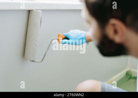 kaukasischer bärtiger Mann, der die Wand unter dem Fenster mit Malerwalze malte. Malerwohnung, renovierendes Haus mit grauer Farbe. Bild mit selektivem Fo Stockfoto
