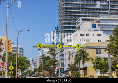 Ein akzentuiertes Schild mit langen Fußgängerwegen hängt über einer der Straßen von Miami Beach mit Hochhäusern und blauem Himmel als Hintergrund. Miami Beach. Stockfoto