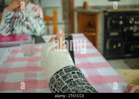 Ein alter Mensch mit einem gegossenen Handgelenk und einem Handy ist in seiner Küche Stockfoto