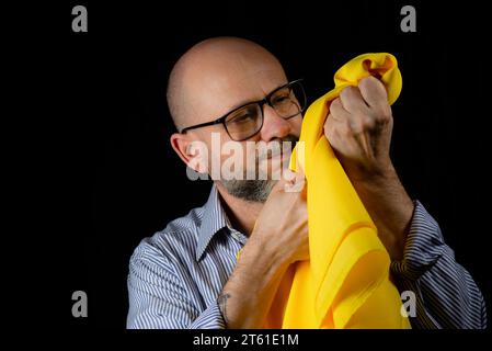 Kahler, bärtiger Mann mit einer Brille, der ein gelbes Tuch über dem Kopf vor schwarzem Hintergrund hält. Stockfoto