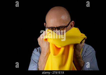 Kahler, bärtiger Mann mit einer Brille, der ein gelbes Tuch über dem Kopf vor schwarzem Hintergrund hält. Stockfoto