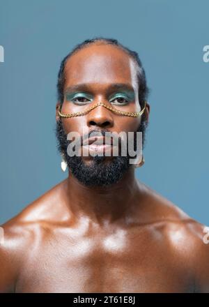 Schwarzer Schwuler steht isoliert auf blauem Hintergrund und starrt mit ernstem Gesichtsausdruck in die Kamera. Nahporträt eines hübschen erwachsenen Transgender mit blauen Lidschatten und goldenem Accessoire im Gesicht Stockfoto