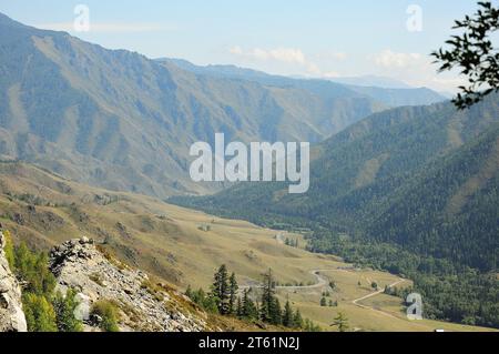 Ein Blick durch die Büsche von einem felsigen Gipfel auf eine gewundene Straße, die an einem sonnigen Herbstabend in ein Tal in den Bergen hinabführt. Chike-Taman-Pass, Stockfoto