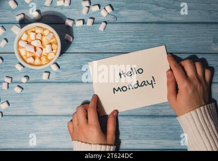 HALLO MONDAY blauer montag Konzepttext weibliche Hände halten Karte mit weißer Tasse Kaffee und Marshmallows auf hölzernem Hintergrund. Der deprimierendste Tag des Jahres im Januar. Tag begehen Selbstmord und Depression Motivationszeichen. Draufsicht, flach Stockfoto