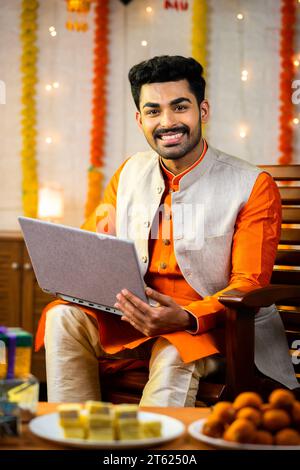Vertikale Aufnahme eines glücklichen jungen indianers, der während des diwali Festivals Laptop benutzt, indem er zu Hause die Kamera betrachtet - Konzept der Festivalfeier, des Verkaufs und Stockfoto
