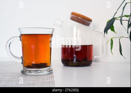 Schwarzer Tee in einer Glasschale und Glas-Teekanne frisch gebrüht auf einer Serviette und grünen Blättern mit weißem Hintergrund Stockfoto