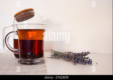 Schwarzer Tee in einer Glasschale und Glas-Teekanne auf einer Serviette und Lavendel auf weißem Tisch mit weißem Hintergrund Stockfoto
