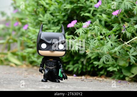 Funko Pop Vinyl Actionfigur des DC-Superhelden Batman Bruce Wayne. Spielzeug steht auf Asphaltstraße im grünen Park, rosa Blumen. Stockfoto