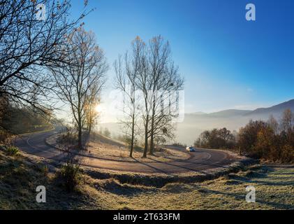 Bergpass im Herbst. Sonne hinter den blattlosen Bäumen auf den grasbewachsenen Hügeln im Frost. Nebel im Tal. Blauer Himmel über dem fernen Kamm Stockfoto