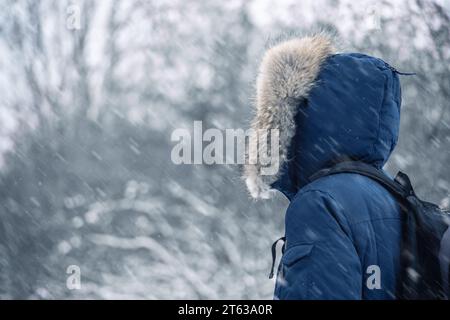 Rückansicht eines Reisenden in einer blauen Jacke mit Pelzhaube und Rucksack vor dem Hintergrund einer Winterlandschaft bei Schneefall Stockfoto