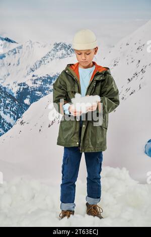 Vertikale Aufnahme eines kleinen Jungen mit Schnee in den Händen in stilvollem, warmem Outfit, Mode Stockfoto