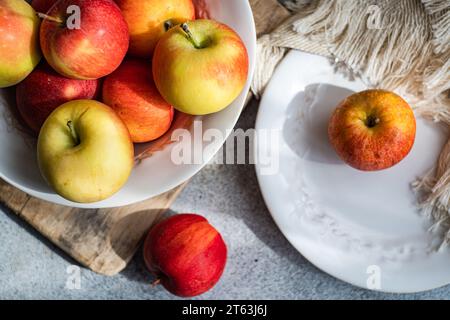 Von unten eine Sammlung reifer, farbenfroher Äpfel, präsentiert auf einem rustikalen Holzbrett, mit einem weißen Teller und drapiertem Stoff, die eine ruhige Küche schaffen Stockfoto