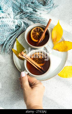 Draufsicht der anonymen Hand, die nach einer von zwei Tassen gewürzten Tees greift, begleitet von Zimtstangen, Anis und getrockneten Orangenscheiben, die in der Nähe von Rad platziert werden Stockfoto