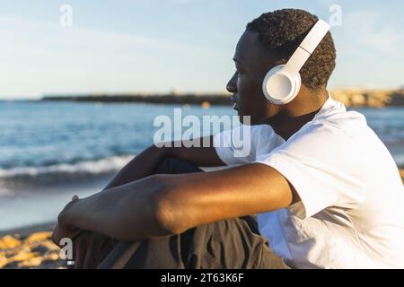 Seitenansicht eines jungen afroamerikanischen Mannes mit Kopfhörern am Strand mit sanften Wellen und weit entfernten Segelbooten im Hintergrund Stockfoto