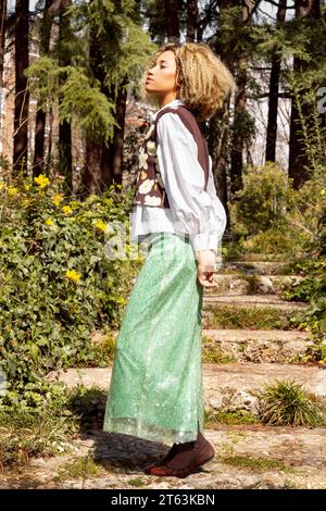 Seitenansicht des ganzen Körpers einer modernen ethnischen Frau mit lockigen Haaren im Paillettenrock, die wegblickt, während sie im Park steht Stockfoto