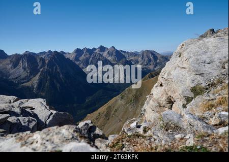 Grasbewachsenes Tal mit Büschen in der Nähe von rauen felsigen Gebirgszügen vor blauem Himmel in der Natur an sonnigen Tagen, Blick vom Tal von Aiguestortes aus Stockfoto