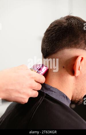 Nahaufnahme der Hand eines Friseurs mit einem violetten elektrischen Clipper auf dem Nacken eines Kunden in einem Salon. Stockfoto