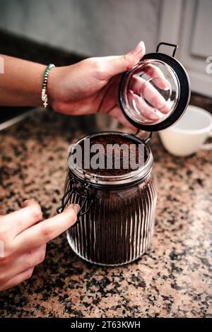 Nahaufnahme einer anonymen Hand, die ein mit fein gemahlenem Kaffee gefülltes Glas öffnet, wodurch die Textur des Kaffees und das Armband am Handgelenk hervorgehoben werden Stockfoto