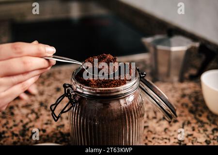 Nahaufnahme einer anonymen Hand, die einen Löffel fein gemahlenen Kaffee aus einem Glas schöpft, mit einer Espressomaschine auf dem Herd und einer Tasse im Hintergrund Stockfoto