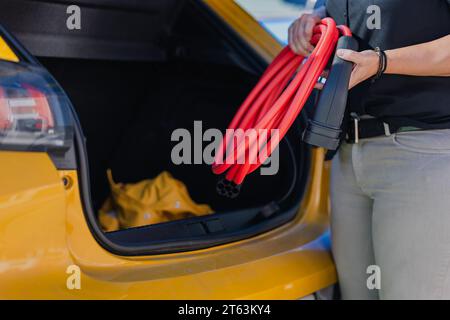 Beschnittene, nicht erkennbare Person, die ein rotes Elektroauto-Ladekabel neben einem geöffneten Kofferraum eines gelben Fahrzeugs hält Stockfoto