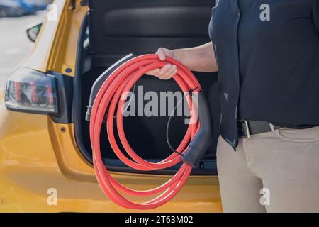 Beschnittene, nicht erkennbare Person, die ein rotes Elektroauto-Ladekabel neben einem geöffneten Kofferraum eines gelben Fahrzeugs hält Stockfoto