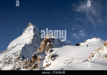 Von unten aus der Rückansicht einer nicht erkennbaren Person mit Skiausrüstung, die an sonnigen Tagen in den Schweizer Alpen auf schneebedeckten Berghängen spaziert Stockfoto