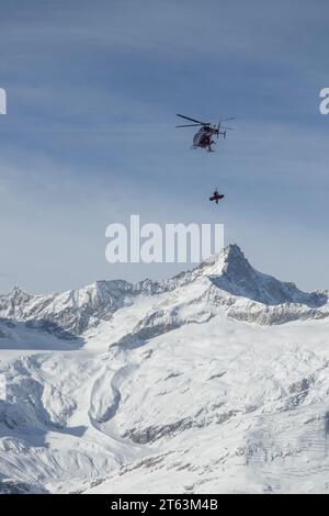 Snowboarder hängt vom Hubschrauber ab, der über schneebedeckte Berge und Landschaft gegen Himmel in den Schweizer Alpen fliegt Stockfoto