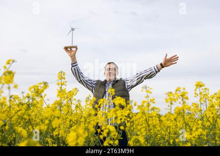 Ein fröhlicher reifer Mann mit Brille hebt ein Windturbinenmodell auf und breitet seine Arme weit in einem gelben Blumenfeld aus. Stockfoto