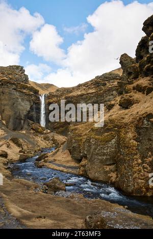 Ein Wasserfall fließt durch zerklüftete Klippen mit einem leuchtend blauen Bach unter einem Himmel mit schwimmenden Wolken, gefangen in Island Stockfoto