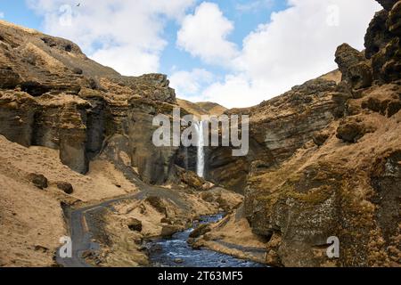 Ein Wasserfall fließt durch zerklüftete Klippen mit einem leuchtend blauen Bach unter einem Himmel mit schwimmenden Wolken, gefangen in Island Stockfoto