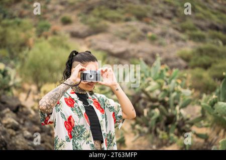 Tätowierte Frau mit lockigen dunklen Haaren in farbenfrohem Hemd, die auf verschwommenem Hintergrund in Masca Teneriffa, den Kanarischen Inseln steht und mit der Filmkamera fotografiert Stockfoto