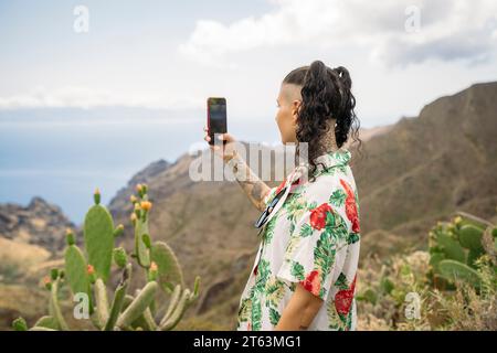 Seitenansicht einer jungen tätowierten Frau in lässiger Kleidung, die Selfie mit Smartphone macht, während sie im Hintergrund der Berge und des bewölkten Himmels in Masca Te steht Stockfoto