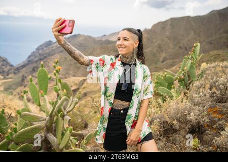 Lächeln einer jungen tätowierten Frau in lässiger Kleidung, die Selfie mit dem Smartphone macht, während sie im Hintergrund der Berge und des bewölkten Himmels in Masca Tene steht Stockfoto