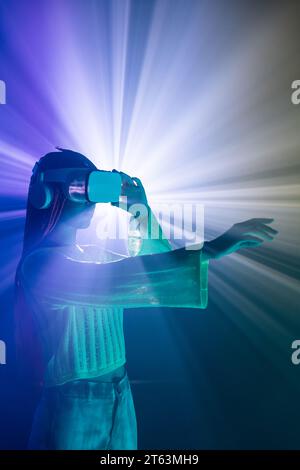 Seitenansicht einer Frau mit orangefarbenen Zöpfen, die eine VR-Brille trägt und die immersive virtuelle Welt über beleuchteten Neonprojektorlichtern erlebt Stockfoto