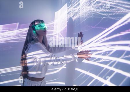 Seitenansicht einer jungen Frau, die futuristische LED-VR-Brille trägt und die Wand mit projizierten Lichtern berührt, während sie den Cyberspace im Studio erkundet Stockfoto