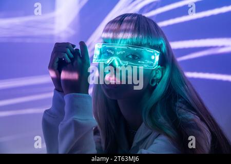 Unemotionale junge Frau, fasziniert von modernen LED-VR-Brillen und eingetaucht in die virtuelle Realität über beleuchteten Projektorlampen Stockfoto