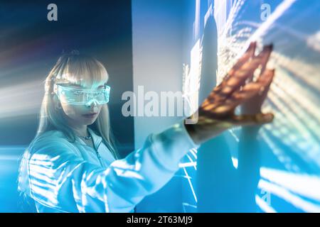Seitenansicht einer jungen Frau, die futuristische LED-VR-Brille trägt und die Wand mit projizierten Lichtern berührt, während sie den Cyberspace im Studio erkundet Stockfoto