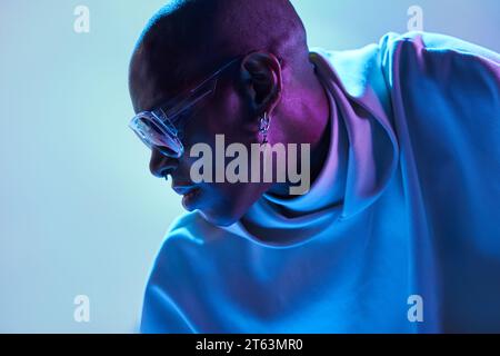 Seriöser afroamerikanischer Glatzkopf mit Ohrring und Sonnenbrille, der in einem modernen Studio mit leuchtenden Lichtern nach unten blickt Stockfoto