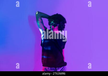 Ein junger lateinischer Mann mit lockigen Haaren schlägt eine dynamische Pose vor einem leuchtend violetten Hintergrund, beleuchtet von Neonlichtern, die von seiner Kleidung reflektieren Stockfoto
