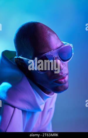 Seriöser afroamerikanischer Glatzkopf mit Ohrring und Sonnenbrille, der in einem modernen Studio mit leuchtenden Lichtern wegblickt Stockfoto