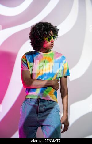 Ein modischer Mann mit lockigen Locken blickt weg und zeigt sein Hemd in Bindefarben vor einem auffälligen Hintergrund mit geometrischem Muster Stockfoto